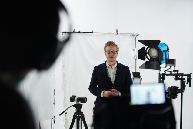 Man wearing a suit filming a video with the help of a teleprompter and recording devices in a studio