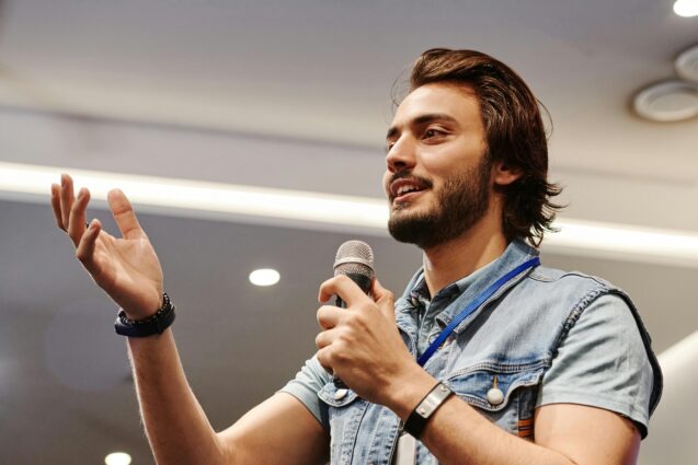 Man holding a microphone while speaking in front of a room full of people