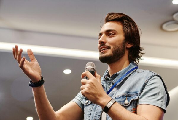 Man with open hand gestures as he holds a microphone with his other hand, and talks to an audience