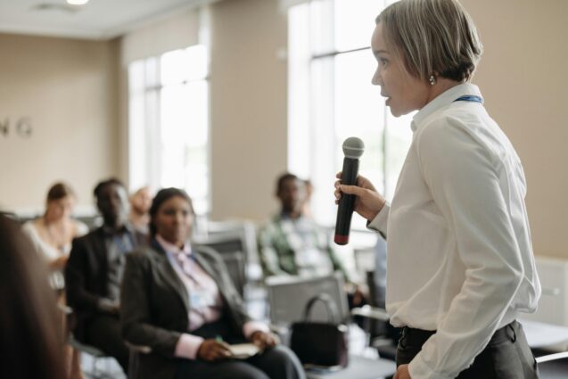 Woman standing in front a room full of people, while holding a microphone