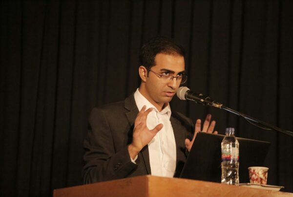 A suited man with glasses addresses an audience, speaking into a microphone