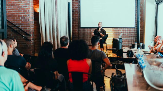 A man sitting while speaking in front of his audience