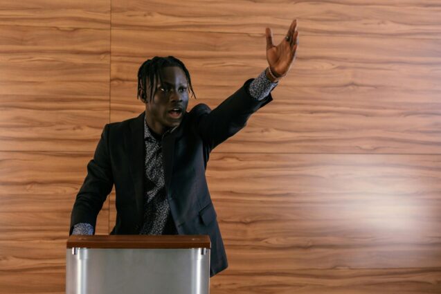 A black man speaking on a podium while raising his other hand