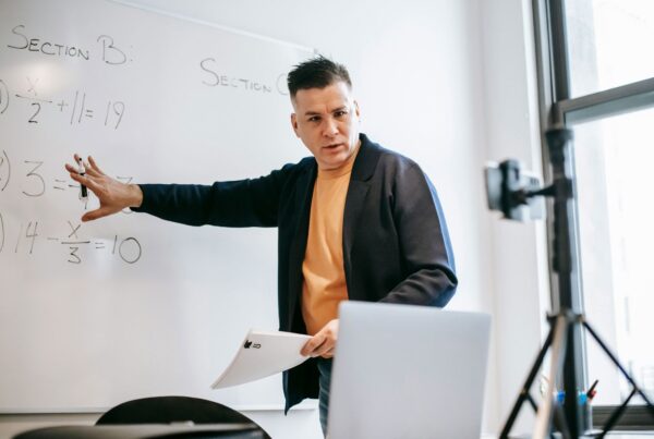 A man is teaching and gesturing in front of a whiteboard while recording a lesson with a camera setup