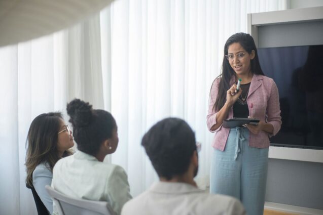 A woman with glasses is leading a meeting, speaking to a group of colleagues while holding a tablet and marker.