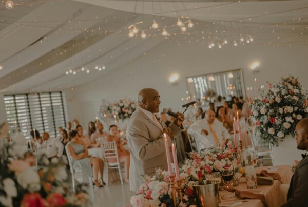 A wedding guest delivers a speech to the audience in front of a beautifully decorated reception room, with floral arrangements and soft lighting