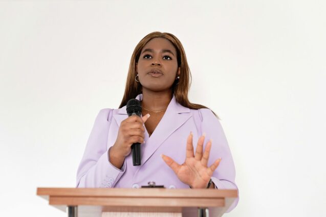 A woman in a lavender blazer speaks into a microphone at a podium