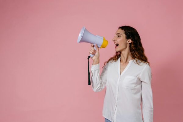 A woman holding a megaphone and shouting against a pink background