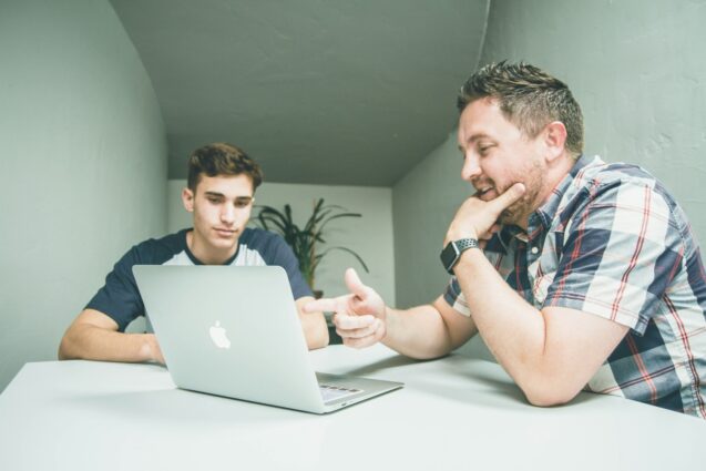 A man and a young man having a discussion in front of a laptop in a modern meeting space.