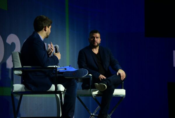 Two men seated on chairs, facing a stage, engaged in conversation