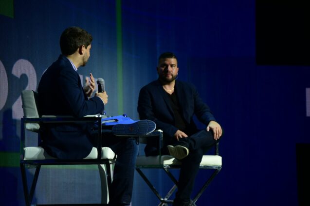 Two men seated on chairs, facing a stage, engaged in conversation