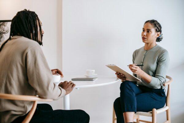 A woman with a notepad listens attentively to a man with dreadlocks during a serious conversation at a white table.