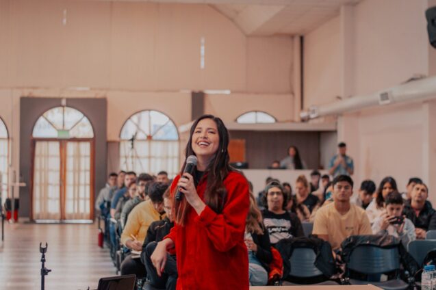 A woman in a red jacket is delivering a speech into a microphone