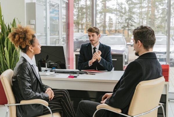 A businessman in a suit speaks to two clients across a desk in a modern office.