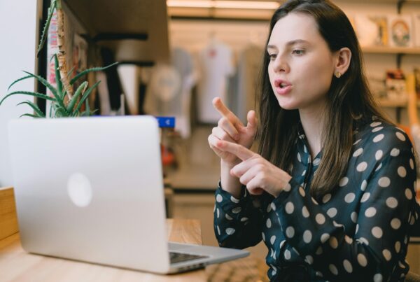 Woman in a polka-dot shirt gesturing while speaking during a video call on her laptop.