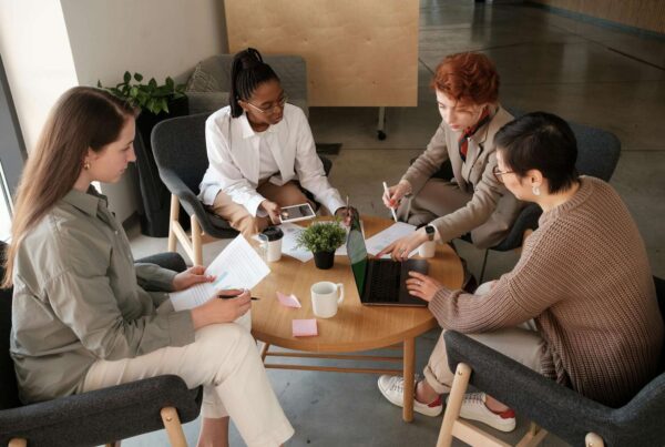 A team of four women collaborating around a table, reviewing documents and using laptops