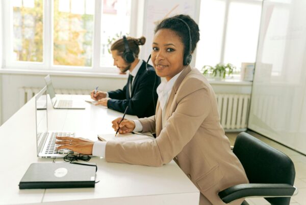 A woman with a headset smiling and writing while a man works on a laptop in a bright office
