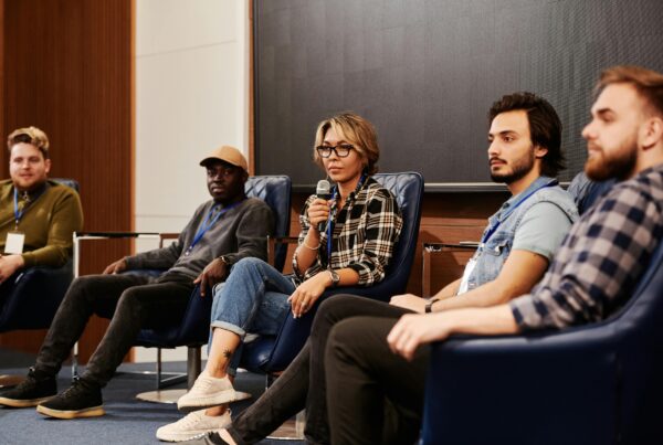 A woman wearing an eyeglasses is making a speech while holding a mic and sitting with other four speakers on stage